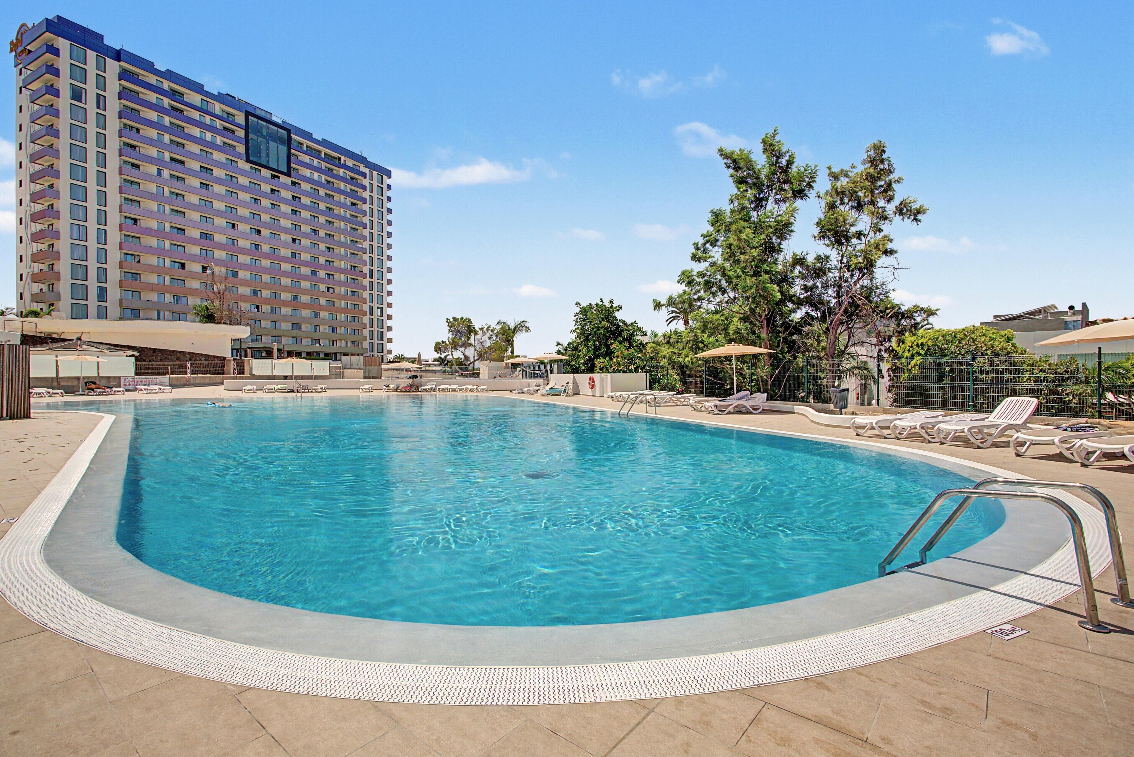 Una piscina con superficie de agua azul cristalina, rodeada de sillones de descanso y un alto edificio de hotel al fondo.