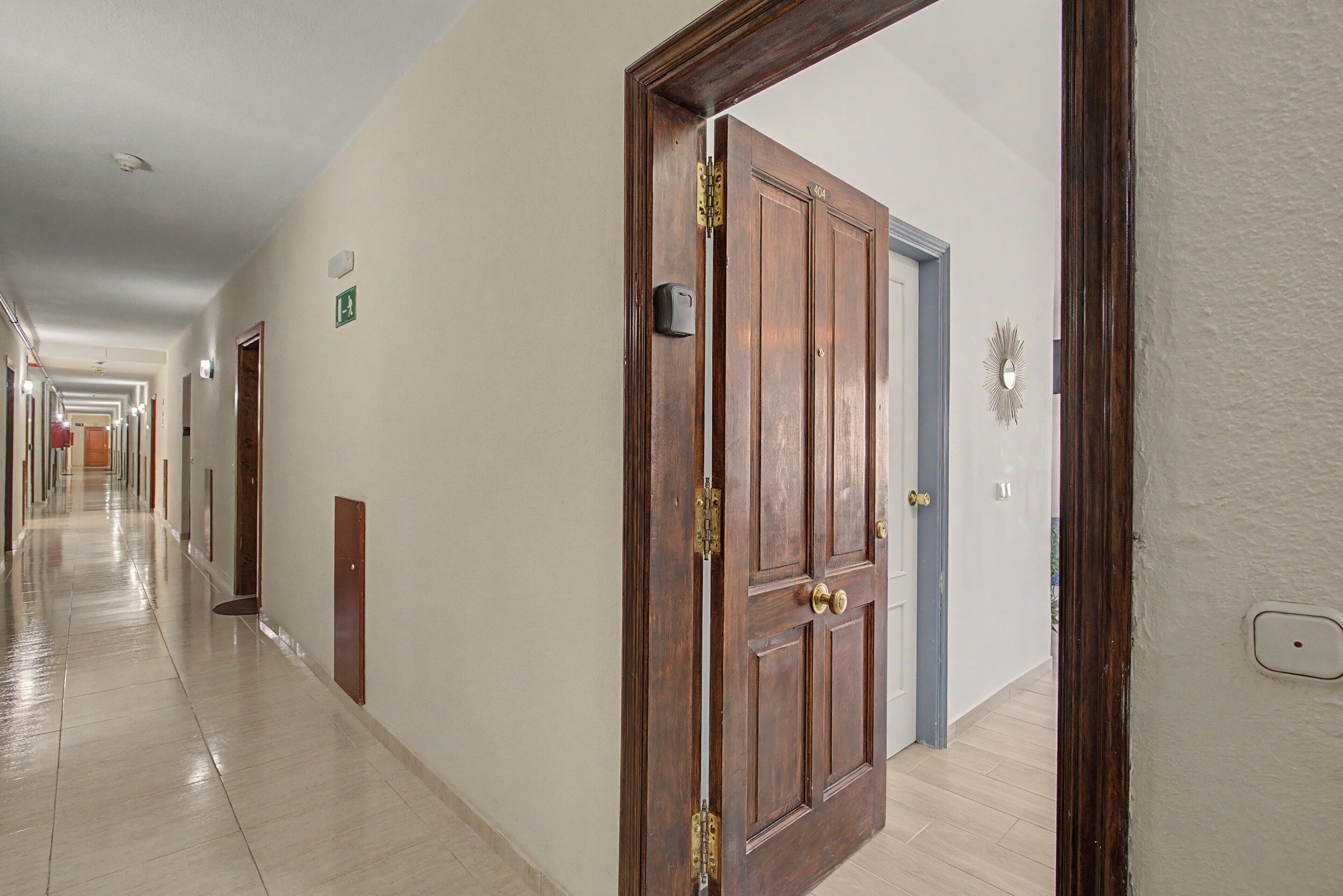 A hotel corridor with wooden doors, a hallway with a tiled floor, and a wall-mounted emergency exit sign.