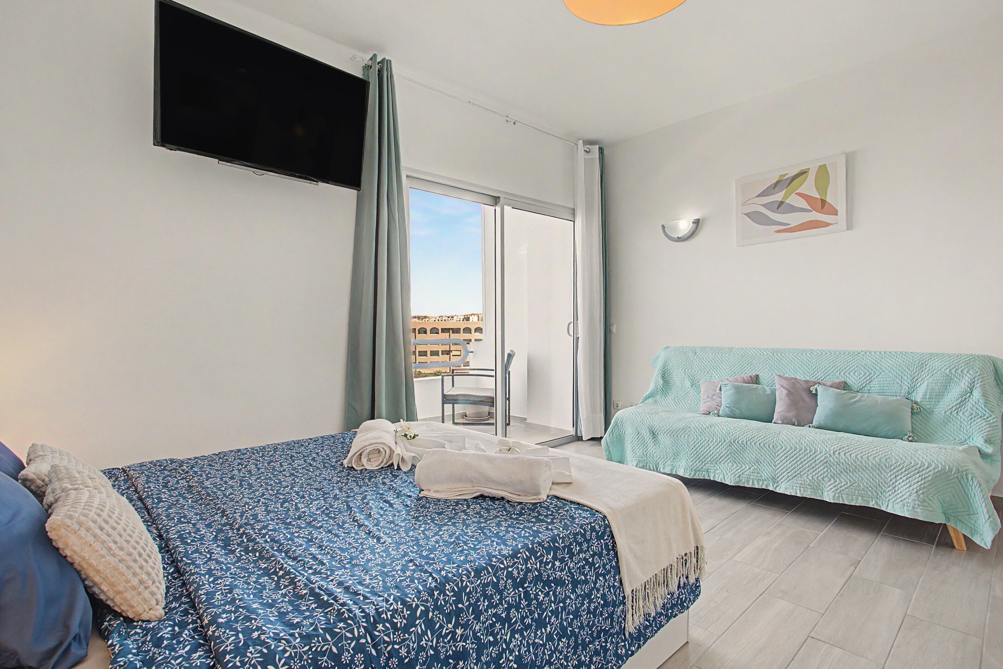 A hotel room with a bed, a sofa, a TV mounted on the wall, and a balcony with a view of buildings.