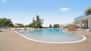 Una piscina con sillones de descanso y sombrillas, rodeada de vegetación y edificios.