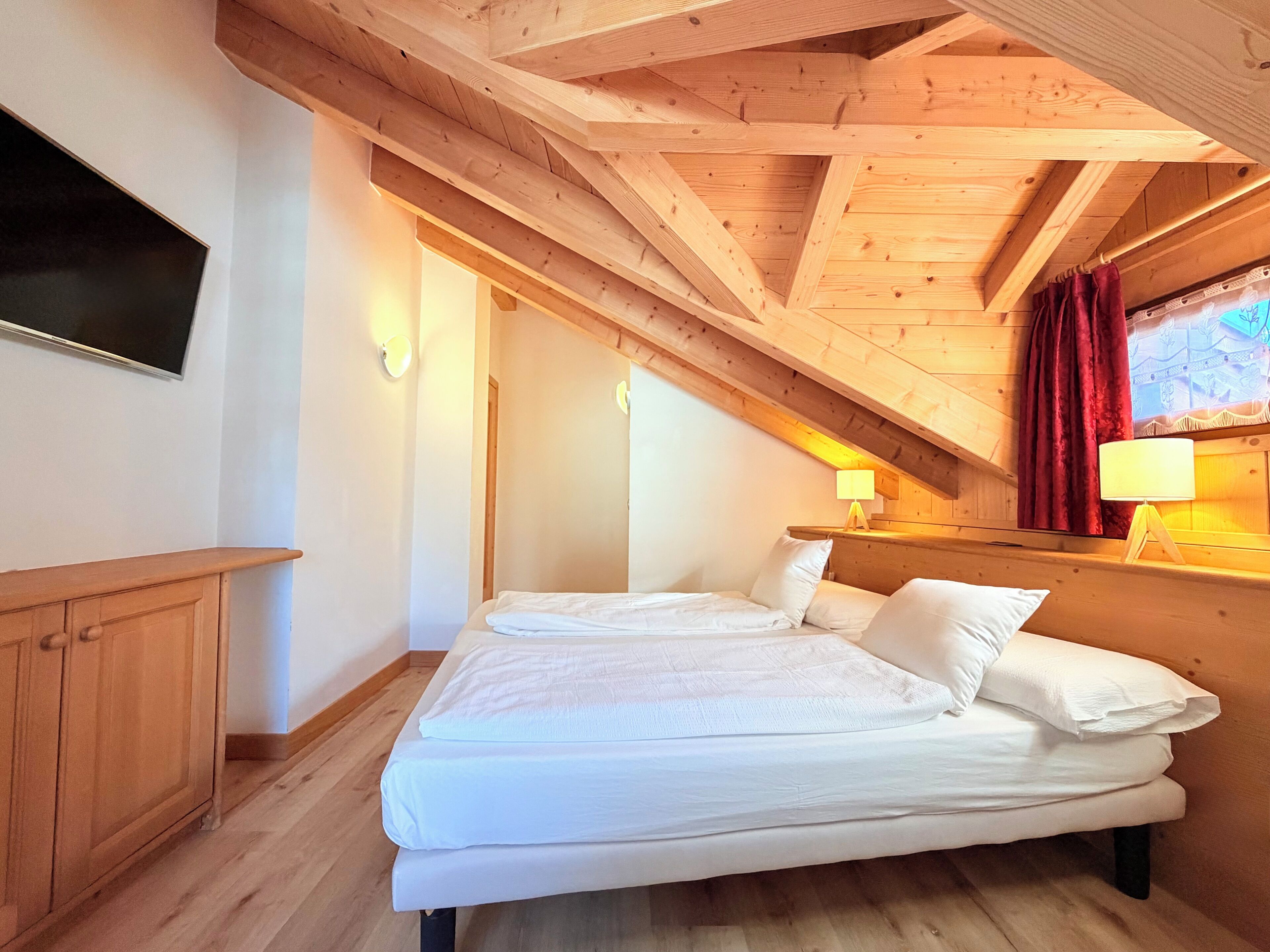 Ein Zimmer mit Bett, Holzbalkendecke, einem an der Wand montierten Fernseher und einem fenster mit Vorhängen.