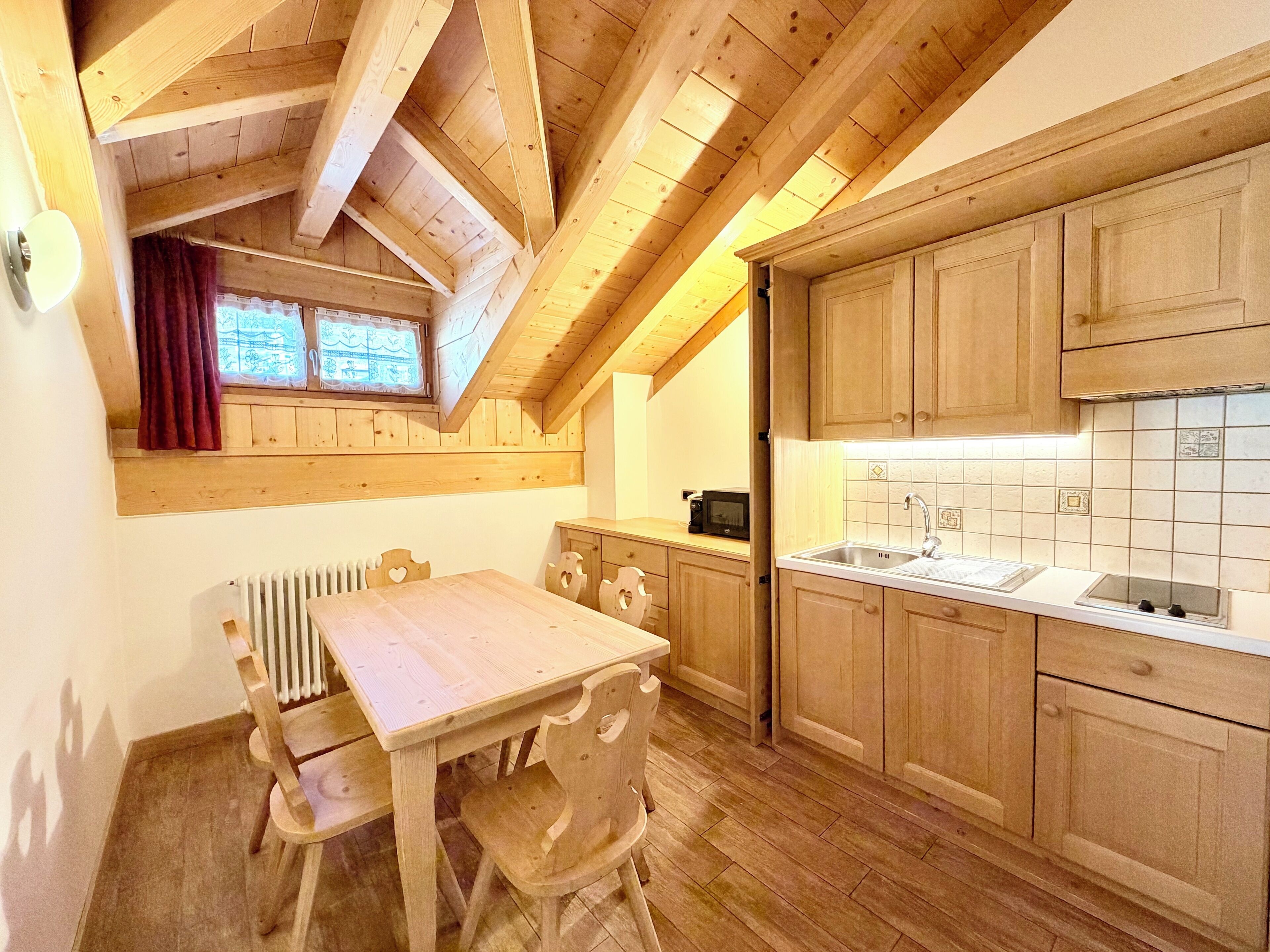 A wooden interior with a dining table and chairs, a kitchen with wooden cabinets, and a window with curtains.