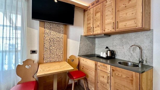 A compact kitchen with wooden cabinets, a built-in TV, a sink, and a small dining area with a table and chairs. - Cesa Planber Apartments Mountain View (Canazei)