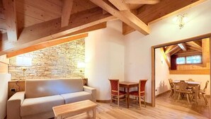 A cozy living room with a stone wall, wooden ceiling beams, a sofa, a coffee table, and a dining area with chairs and a table. - Cesa Planber Apartments Mountain View (Canazei)