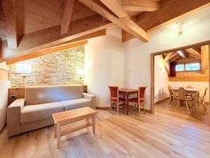 A cozy living room with a stone wall, wooden ceiling beams, a sofa, a coffee table, and a dining area with chairs and a table. - Cesa Planber Apartments Mountain View (Canazei)