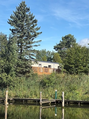 A cabin with a deck, surrounded by dense trees and a calm body of water.