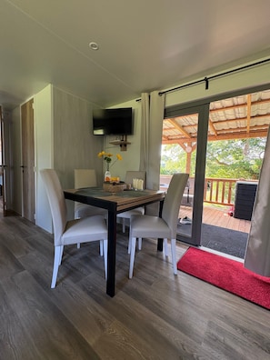 A modern dining area with a table and chairs, a flat-screen TV mounted on the wall, and a view of the outdoors through a glass door.