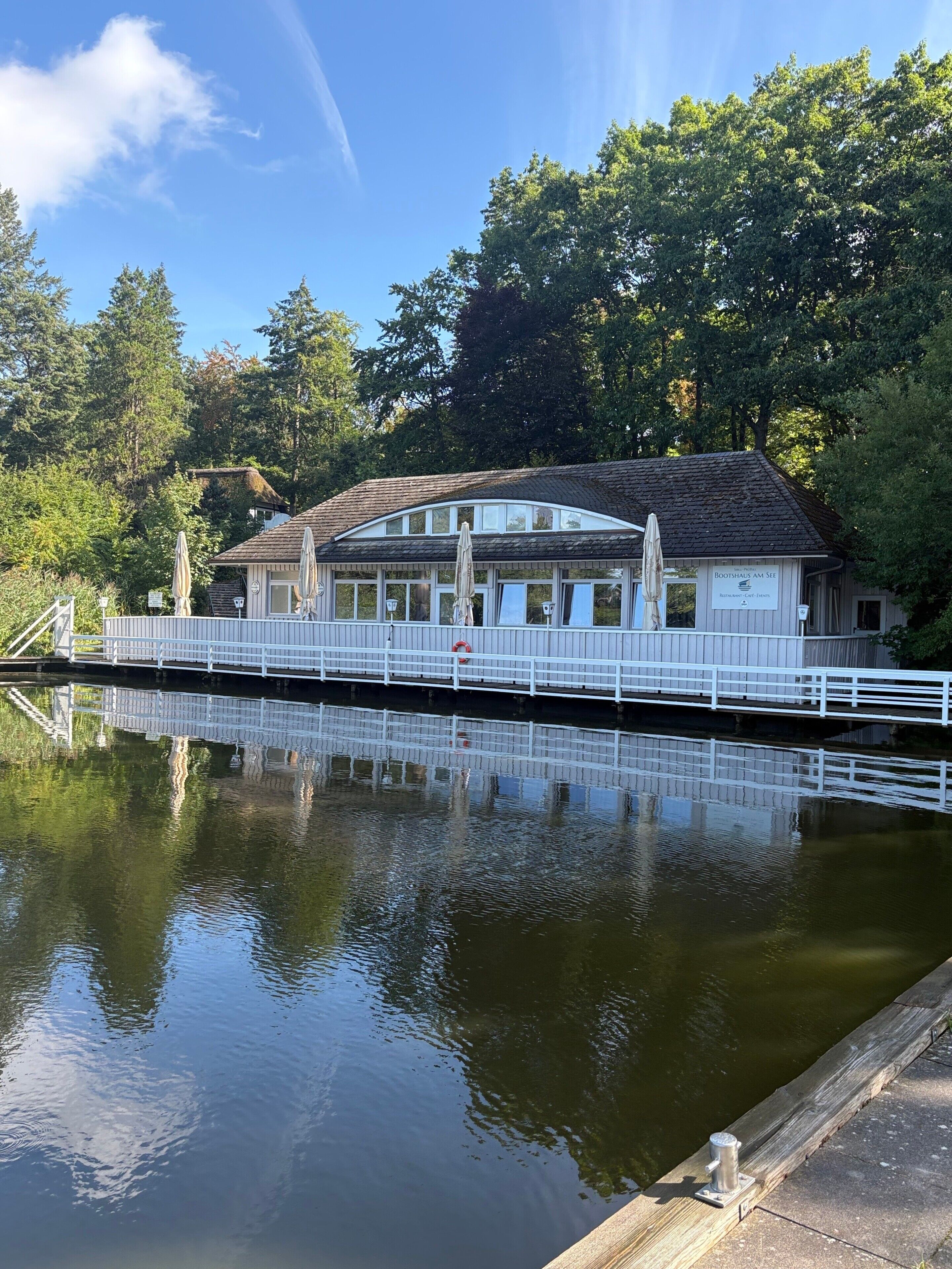 En bygning med et skilt, der viser 'Bootshead's Am See' foran, et hvidt hegn rundt om ejendommen og et roligt vandareal i forgrunden.