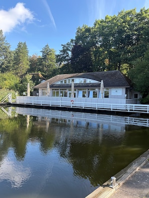 A building with a sign reading 'Bootshead's Am See' across the front, a white fence surrounding the property, and a calm body of water in the foreground.