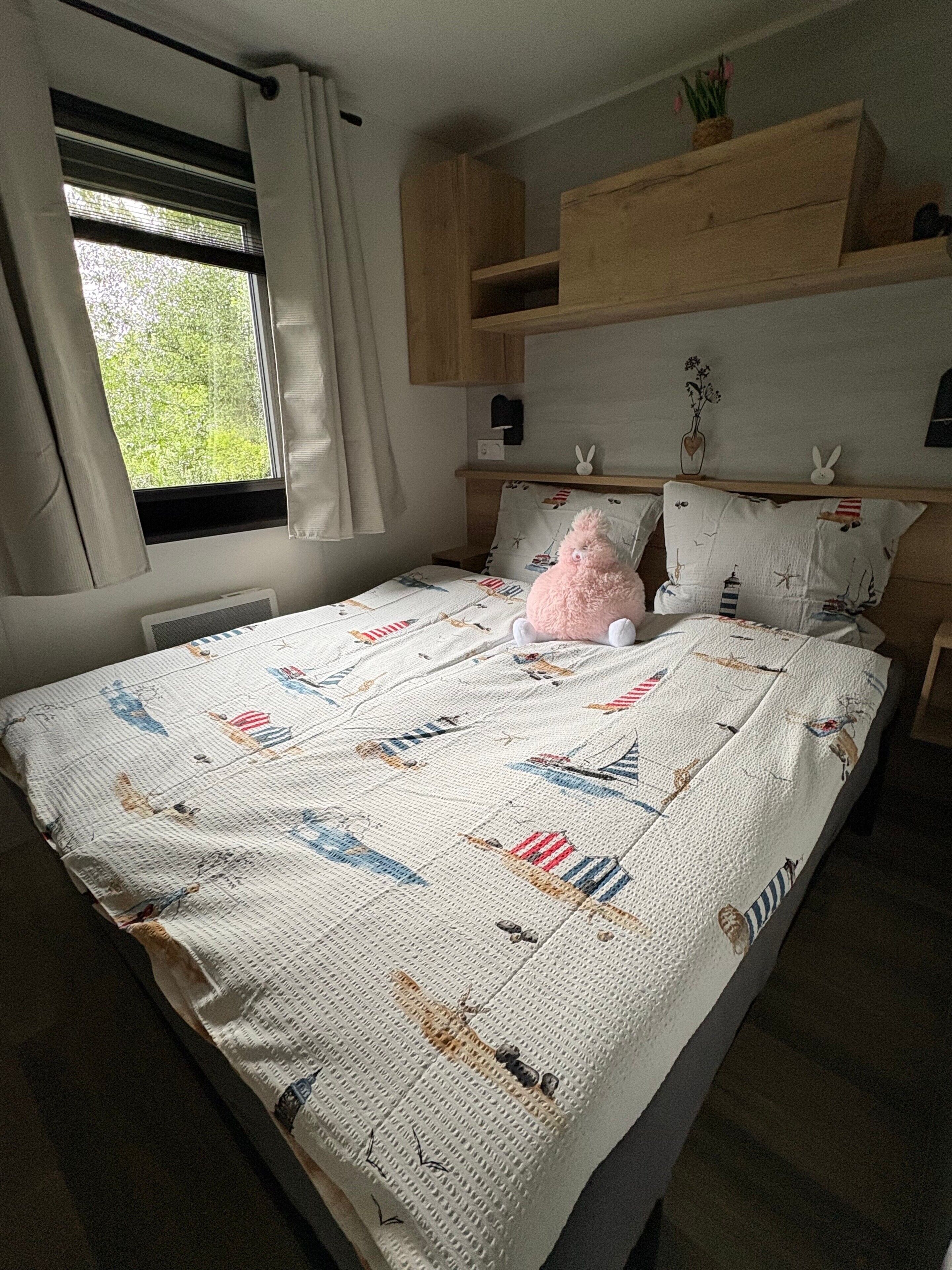 Ein Schlafzimmer mit einem Bett, einem Fenster mit Vorhängen, einem Nachttisch und einem an der Wand befestigten Regal mit Dekoration.