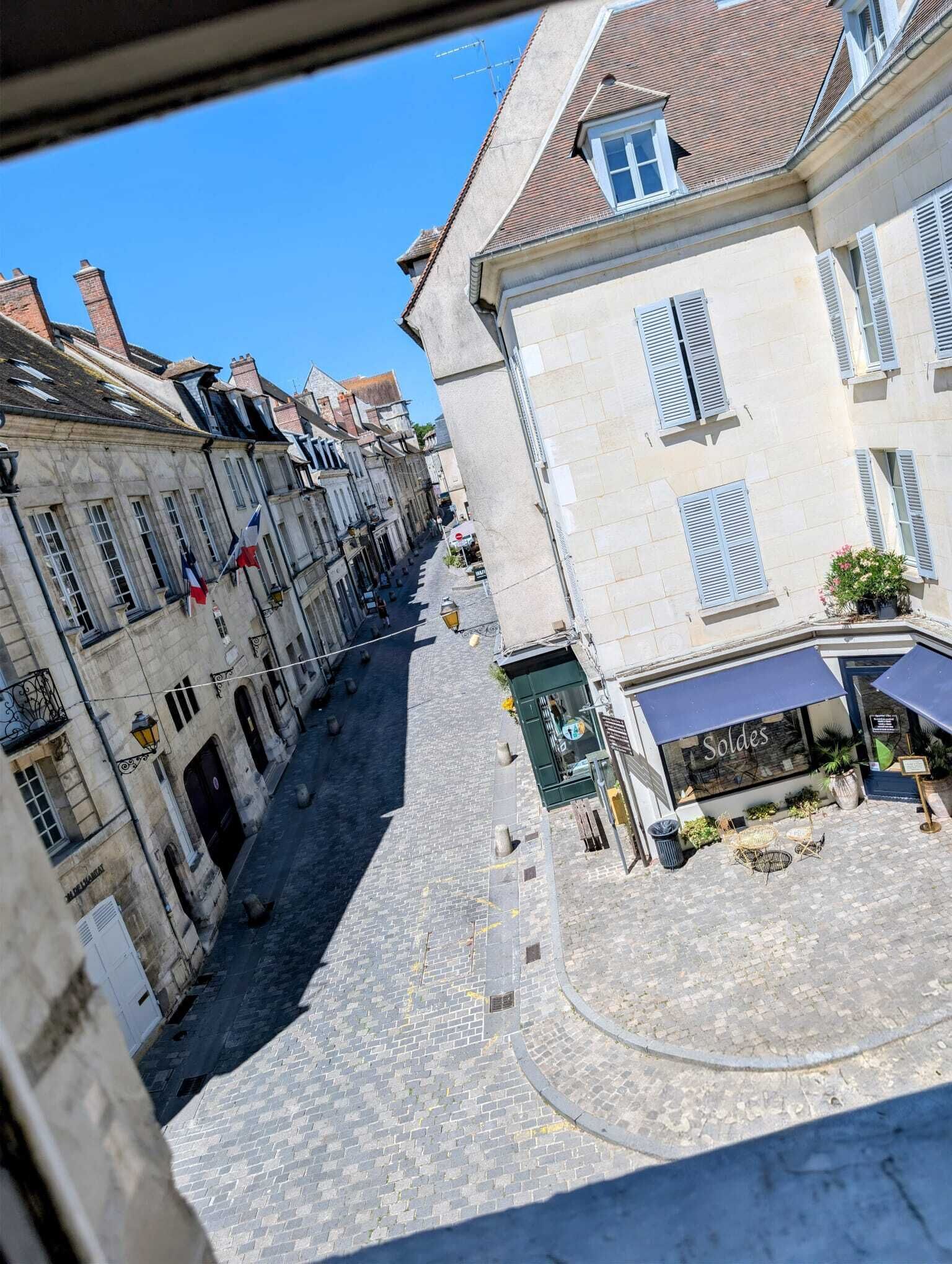 Une ruelle pavée bordée de bâtiments historiques, un café appelé « Soldes » et un ciel d’un bleu limpide.