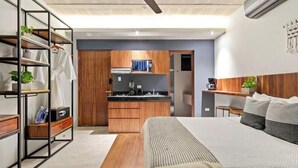 A modern bedroom with a large bed, a wooden wardrobe, a built-in kitchen, and a shelving unit with plants. - Ocean Boutique Condos (Playa del Carmen)