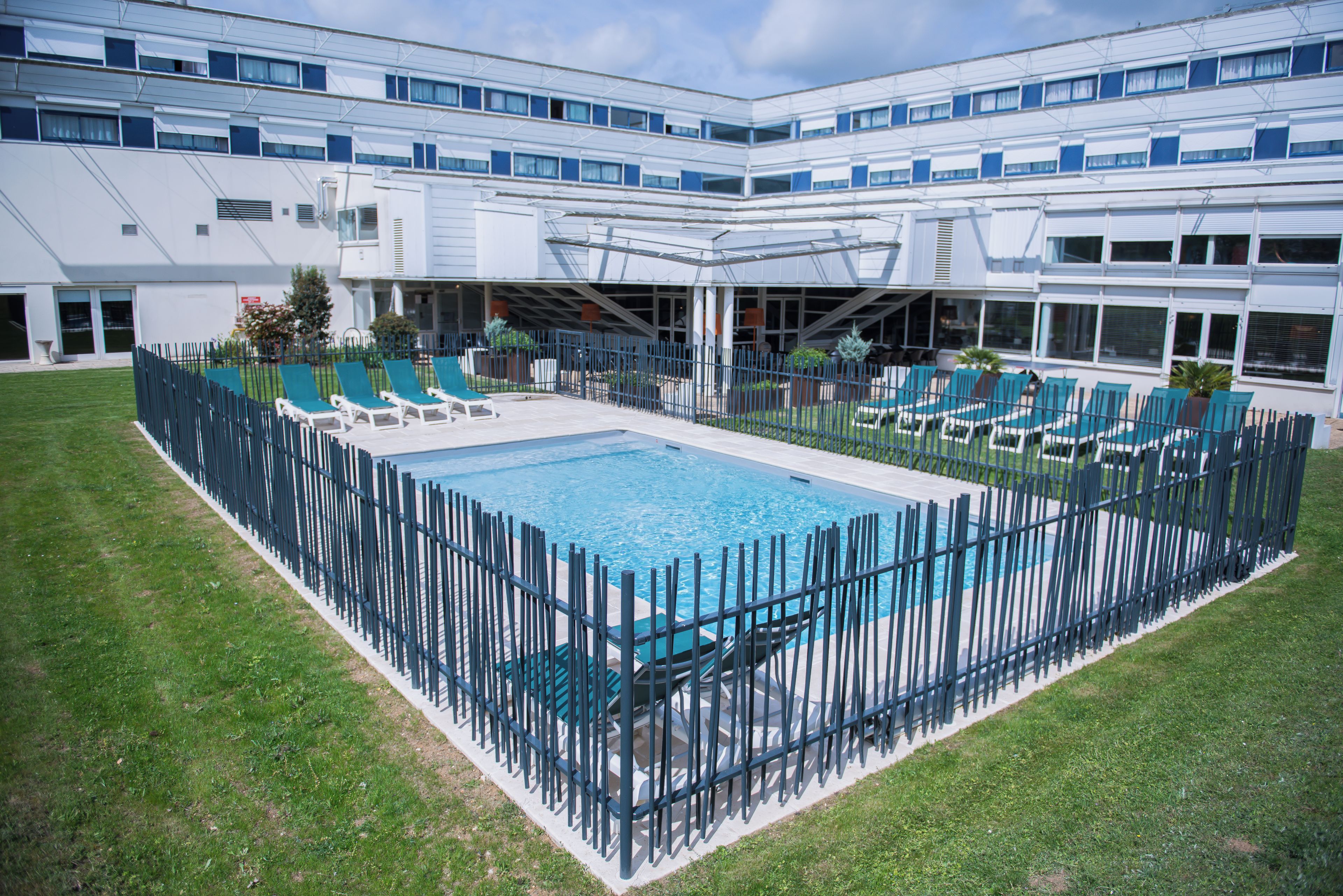 Une piscine entourée d’une clôture, avec des chaises longues et un bâtiment en arrière-plan.