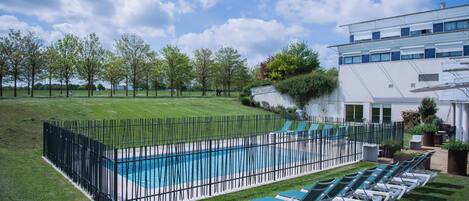 Una piscina rodeada de césped y árboles, con una cerca negra. Se ven sillas de descanso y un edificio con ventanas al fondo.