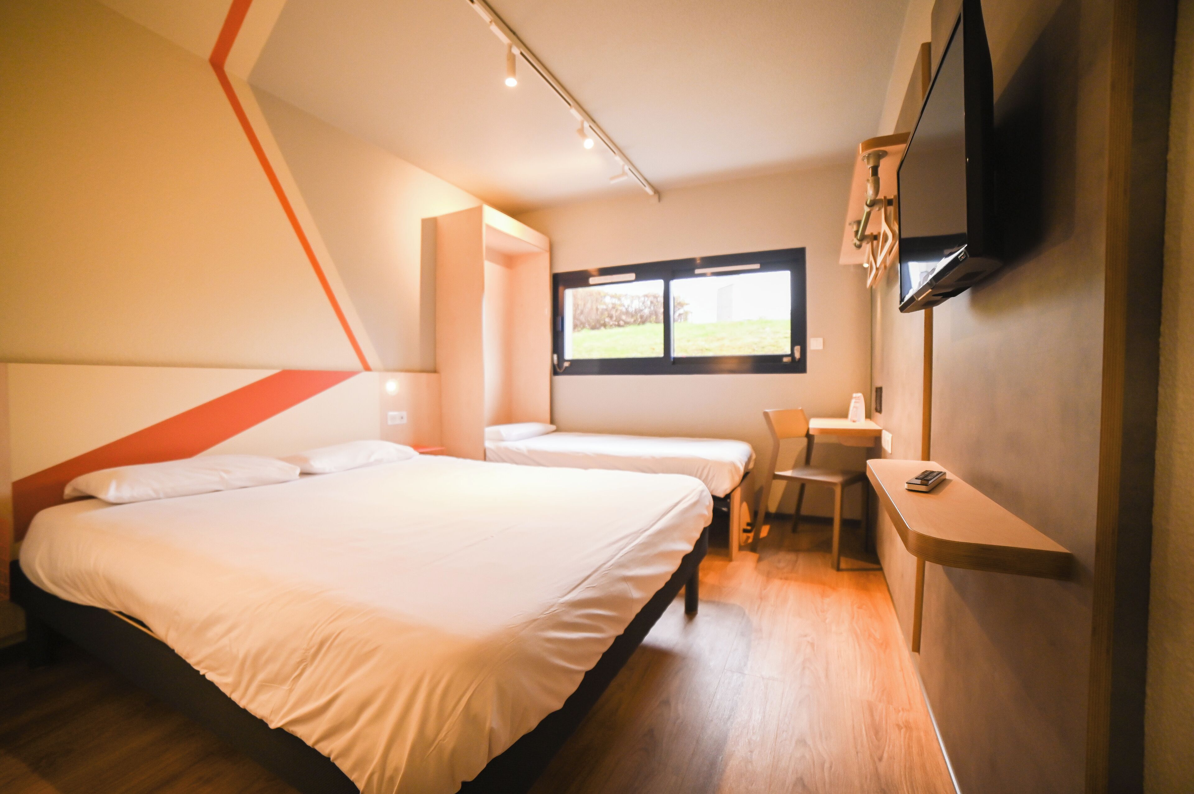 Habitación de hotel con dos camas, un escritorio, una silla, un televisor montado en la pared y una ventana con vistas al exterior.