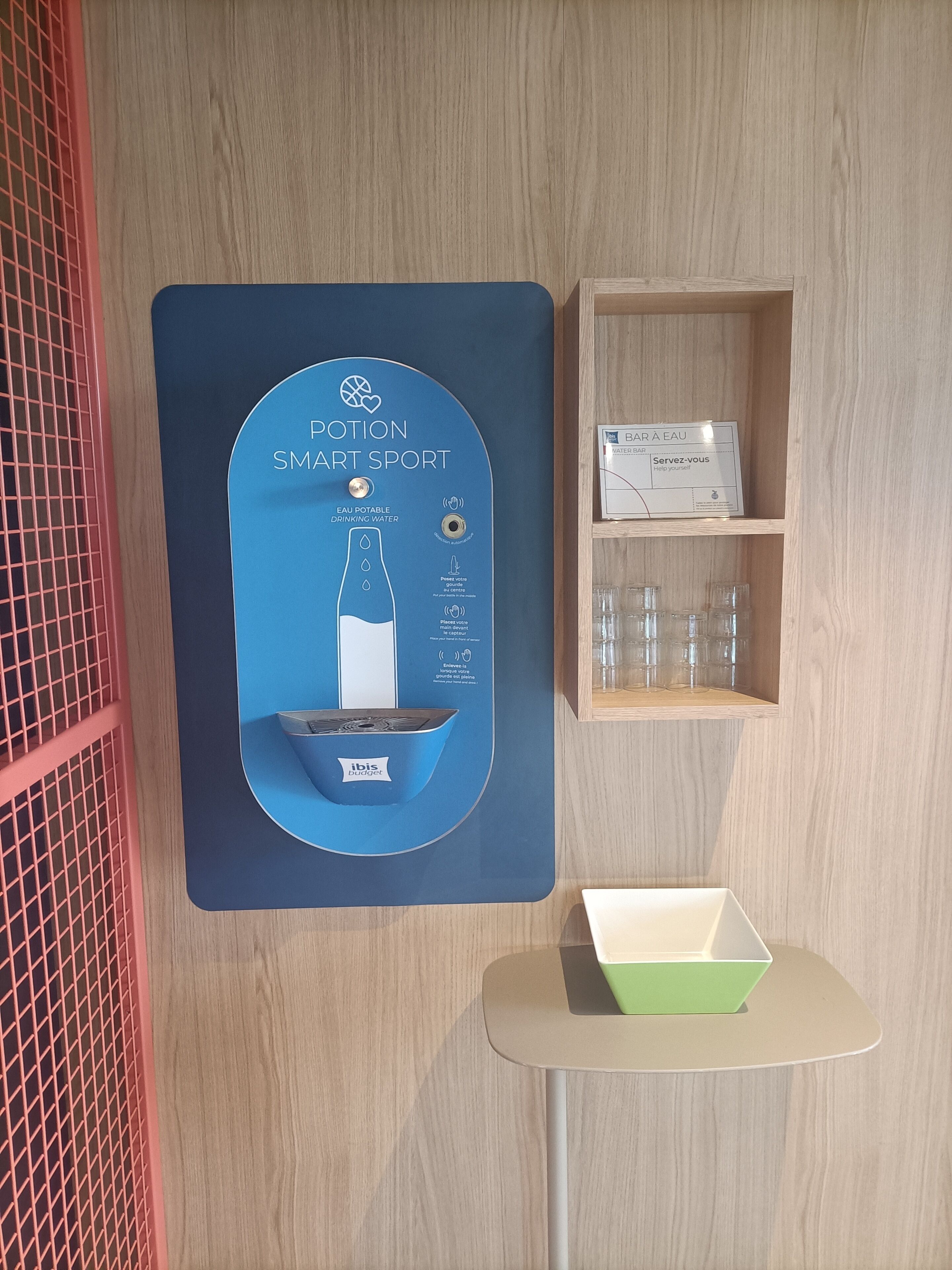 A room with a blue wall-mounted dispenser for drinking water, a small shelf with glasses, and a table with a green box.
