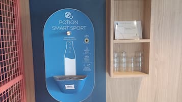 A room with a blue wall-mounted dispenser for drinking water, a small shelf with glasses, and a table with a green box.