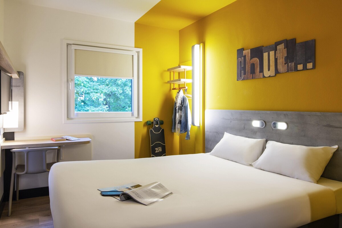 A hotel room with a bed, a desk, a chair, a window, and a wall with a sign.