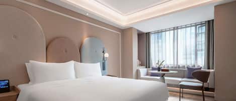 A modern hotel room with a large bed, a seating area with a table, and a large window with curtains.