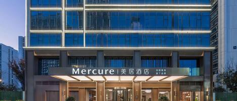 A modern hotel with a glass facade, a well-lit entrance, and a landscaped area with flower beds.