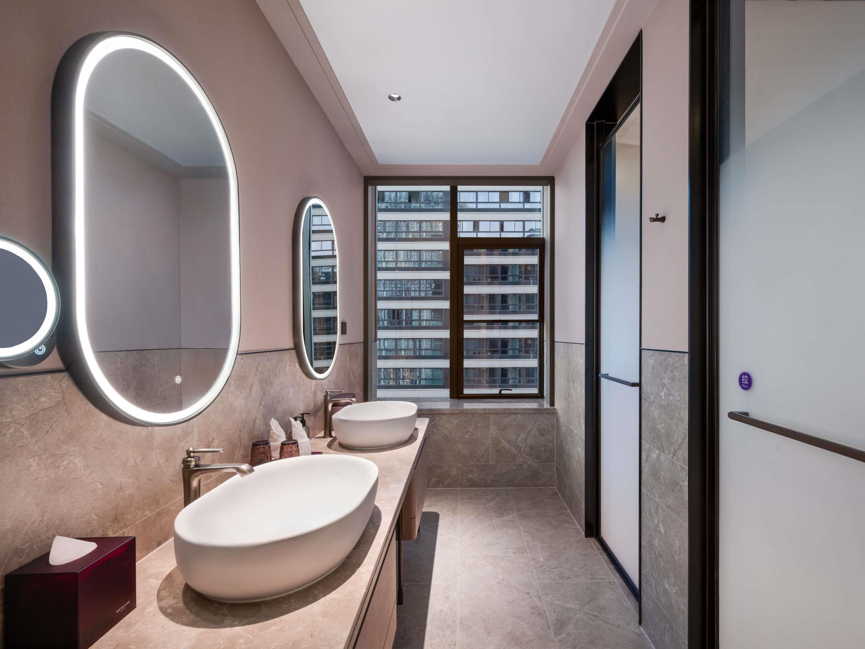 A modern bathroom with two sinks, a large mirror, and a view of a building through the window.