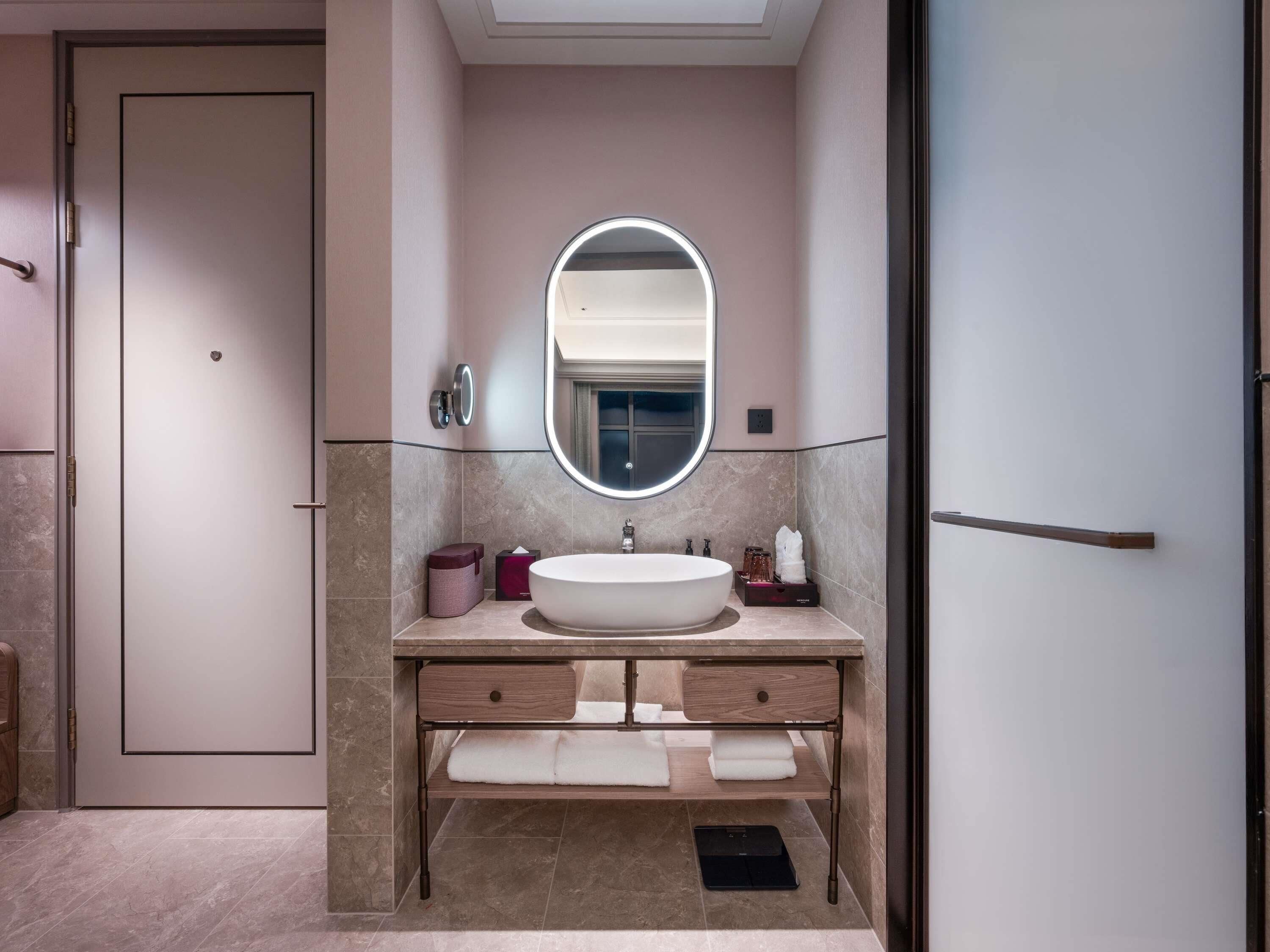 Une salle de bain moderne avec un grand miroir ovale, un lavabo blanc et un meuble-lavabo en bois doté d’un porte-serviettes.