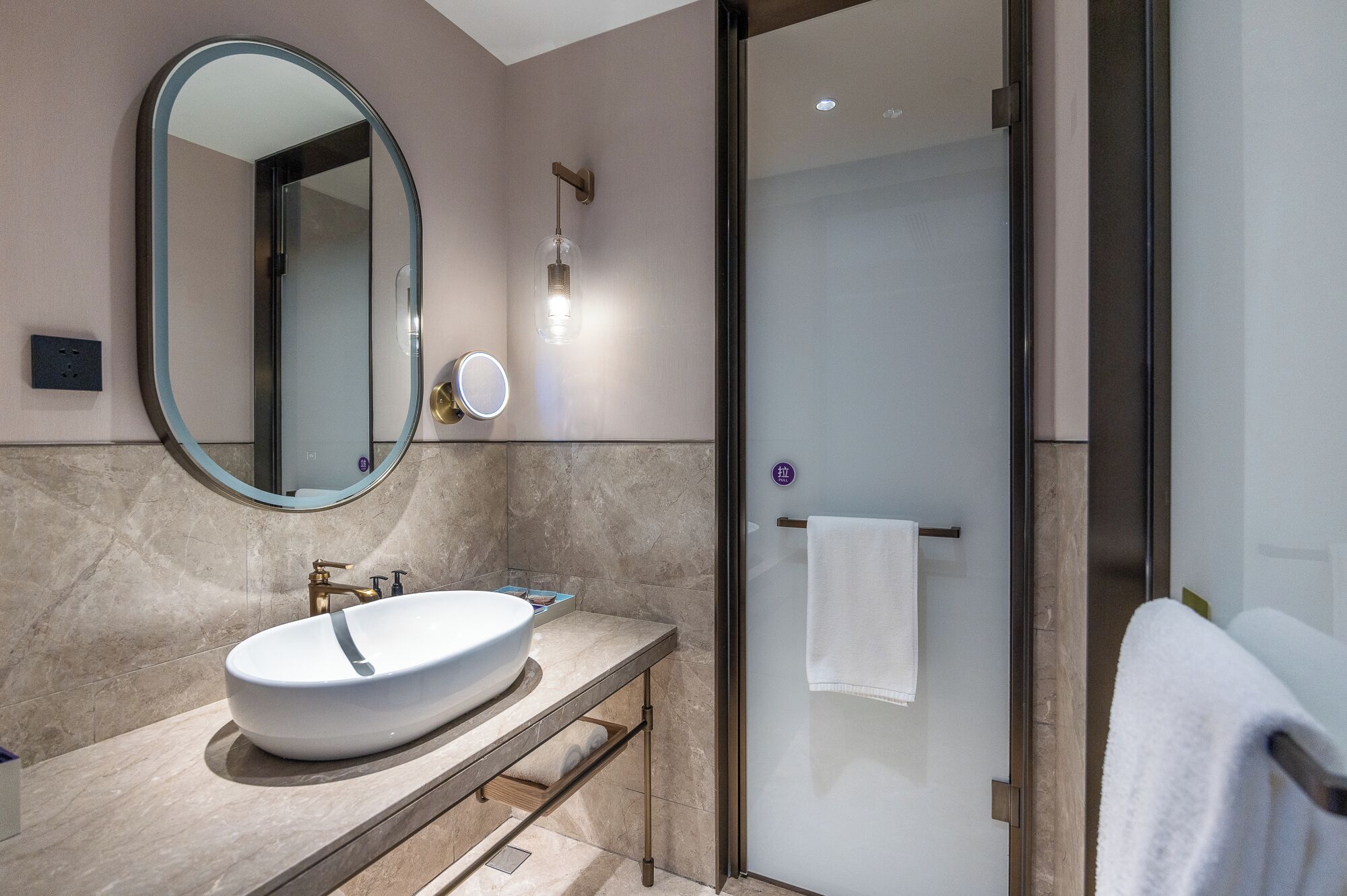 A modern bathroom with a large oval mirror, a white sink, and a glass-enclosed shower.