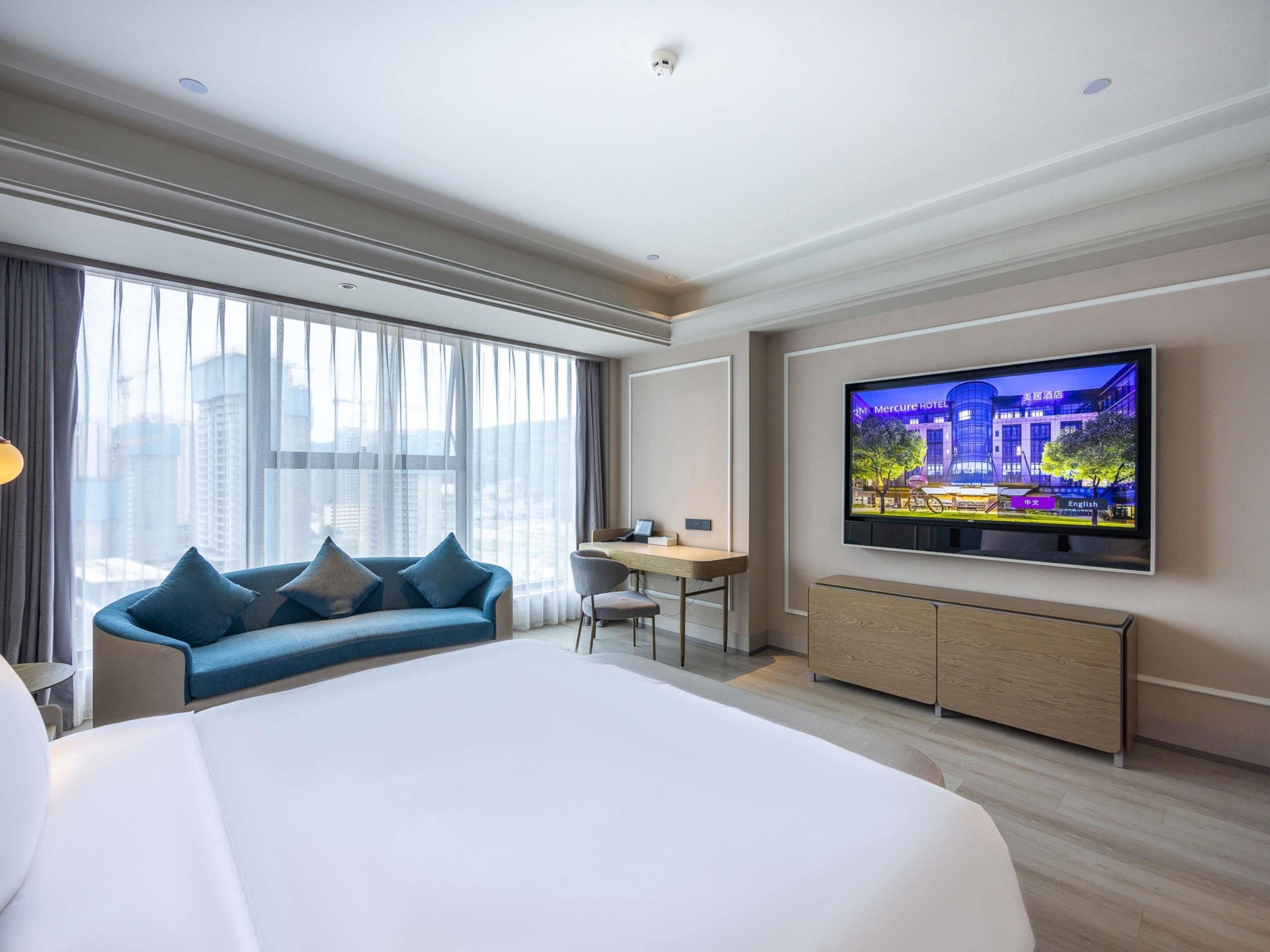 Habitación de hotel moderna con una cama grande, un escritorio, televisión y vistas de la ciudad a través de grandes ventanales.