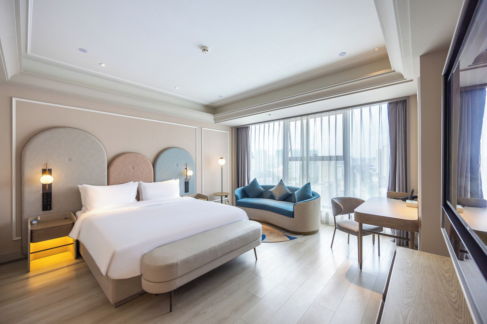 Une chambre d’hôtel moderne dotée d’un grand lit, d’un canapé, d’une table à manger et offrant une vue sur la ville grâce à de grandes fenêtres.