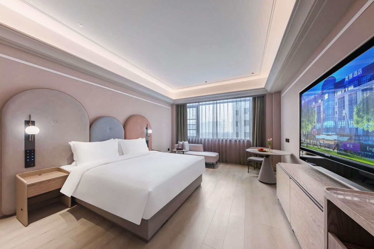 Habitación de hotel moderna con una cama grande, mesitas de noche, zona de estar, televisor y vistas del exterior del hotel.