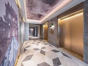 Un pasillo con piso de diseño geométrico, un mural en la pared y un moderno ascensor con una lámpara fijada a la pared.
