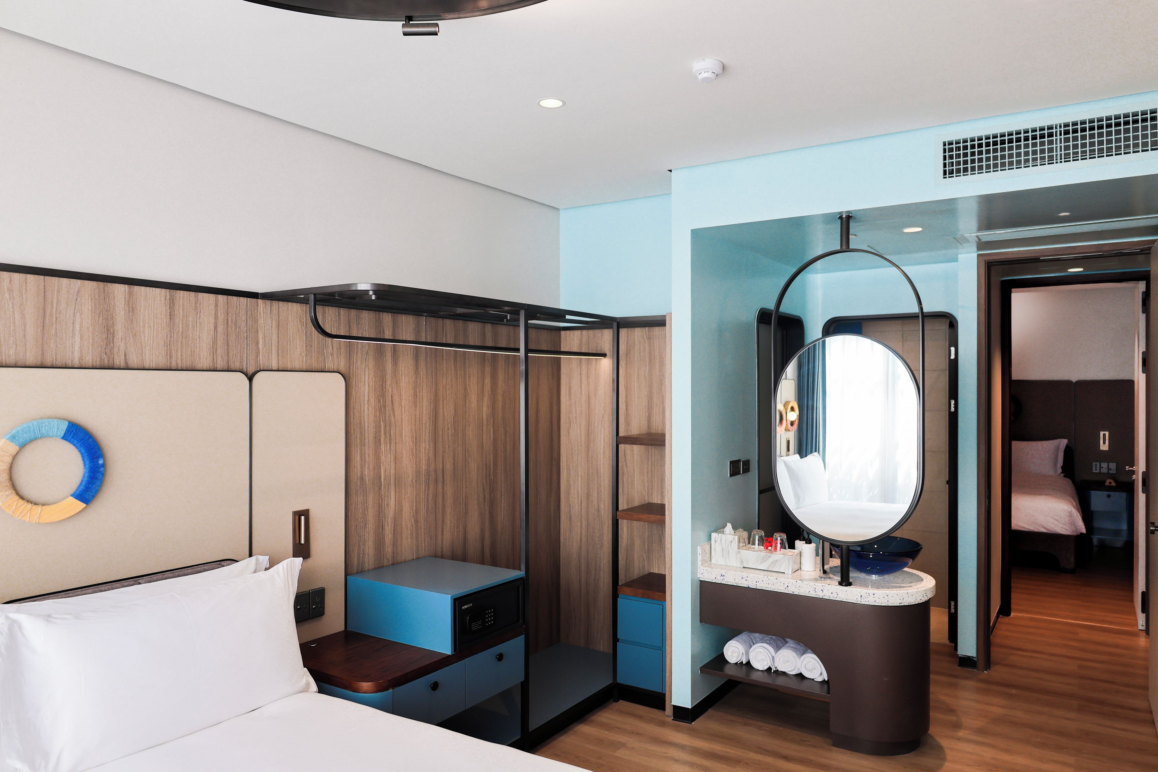 A modern hotel room with a bed, a vanity with a mirror, a walk-in closet, and a bathroom visible through an open door.