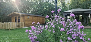 Eine Holzhütte mit einem eingezäunten Garten, einem Pavillon und einem Beet lila Blumen im Vordergrund.