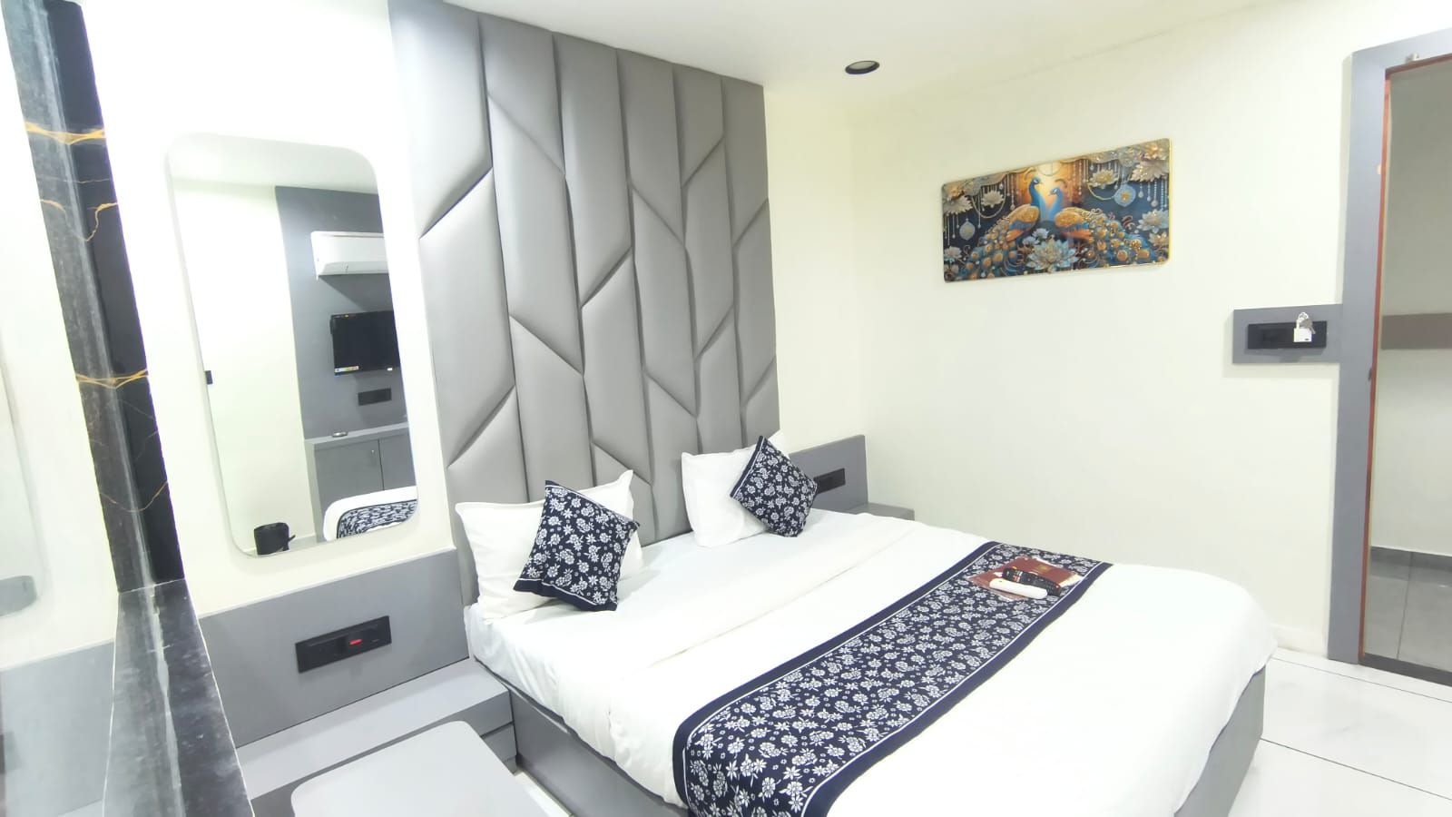 A modern hotel room with a large bed, a built-in headboard, a bedside table, a mirror, and a decorative wall piece.
