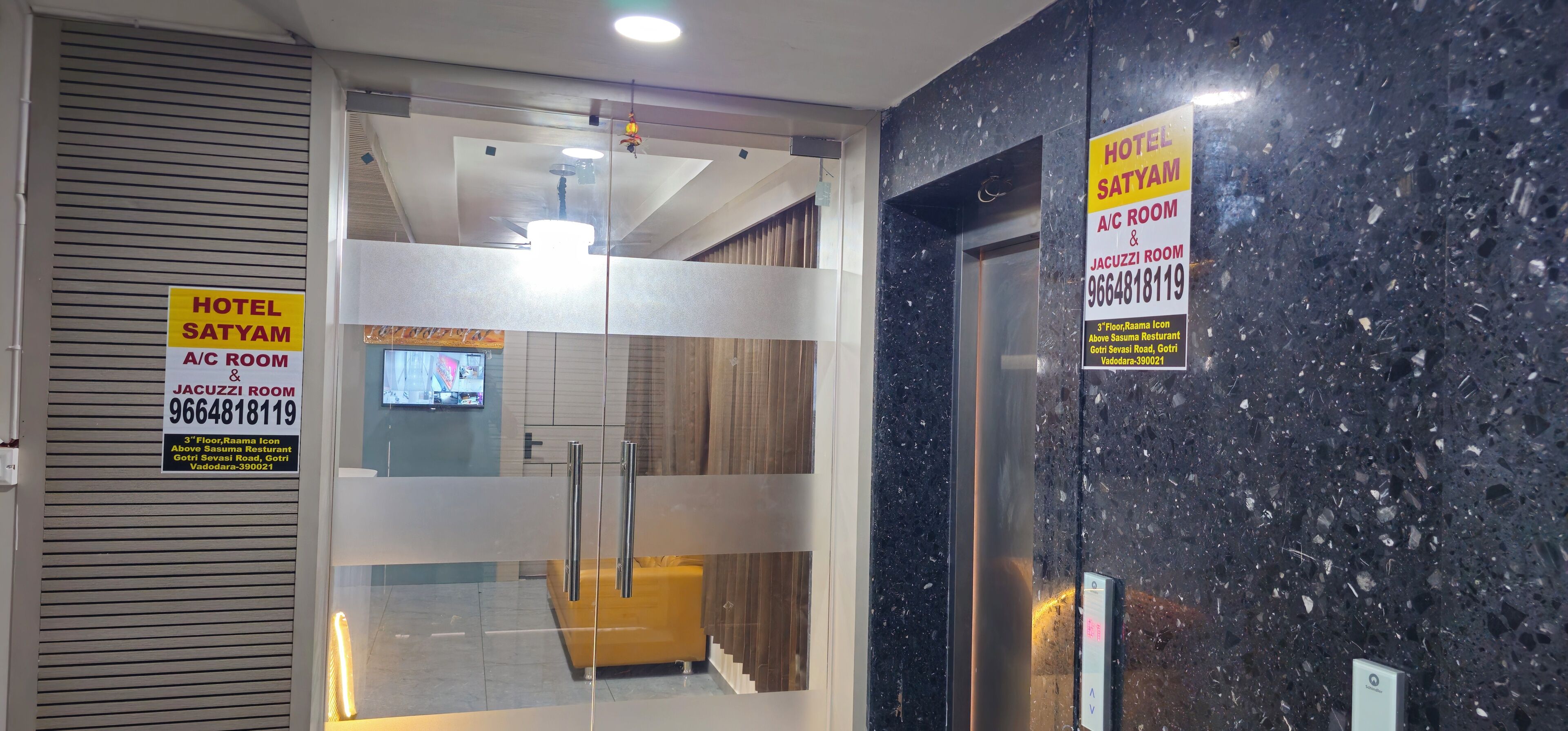 A hotel entrance with a glass door, a signboard with room details, and a marble wall.