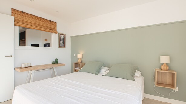 Quarto com cama, mesas de cabeceira, escrivaninha, espelho e prateleira na parede.