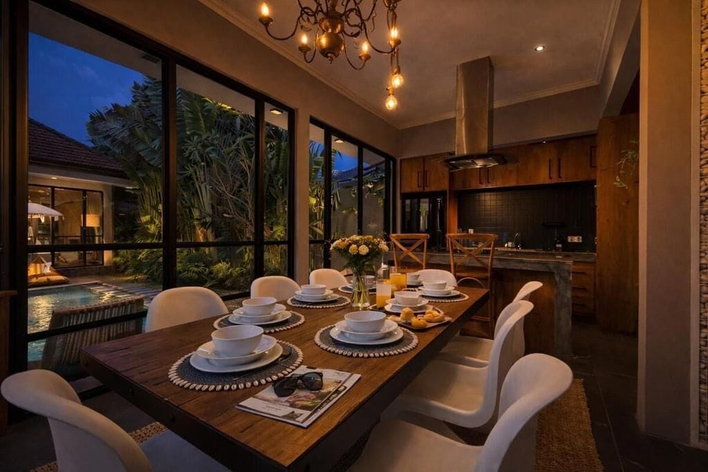 Comedor con una mesa larga dispuesta para una comida, ventanas amplias con vistas a una piscina y una cocina visible al fondo.