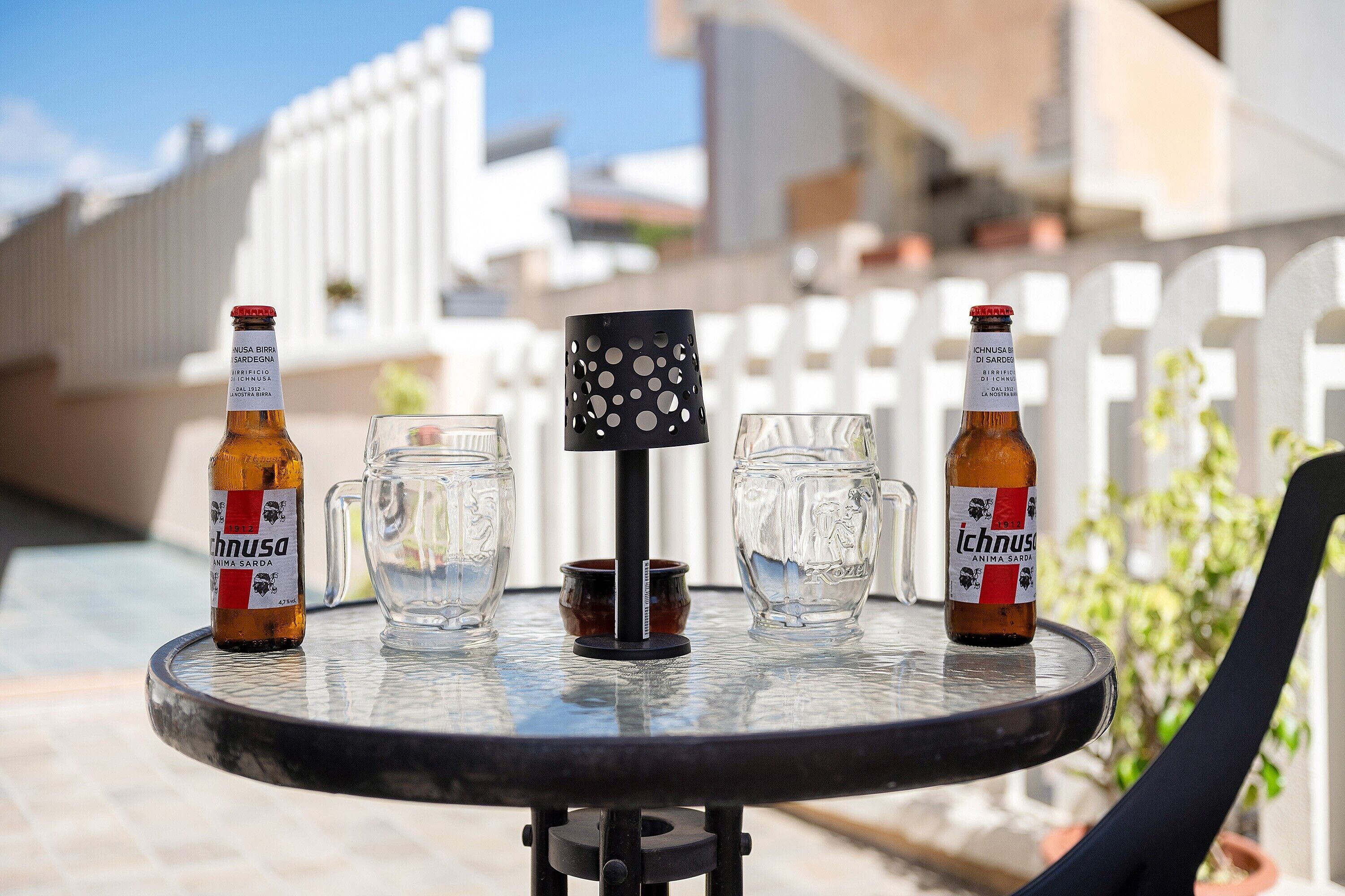 Una mesa redonda con dos botellas de cerveza, dos vasos y un posavasos negro con agujeros.
