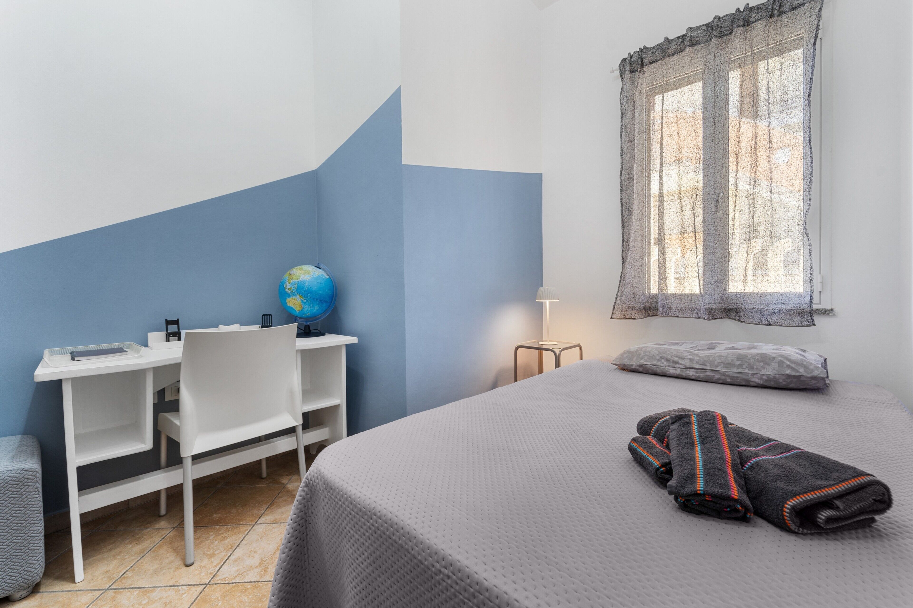 Ein Schlafzimmer mit einem Bett, einem Schreibtisch mit Globus, einem Stuhl, einer Lampe und einem Fenster mit Vorhang.