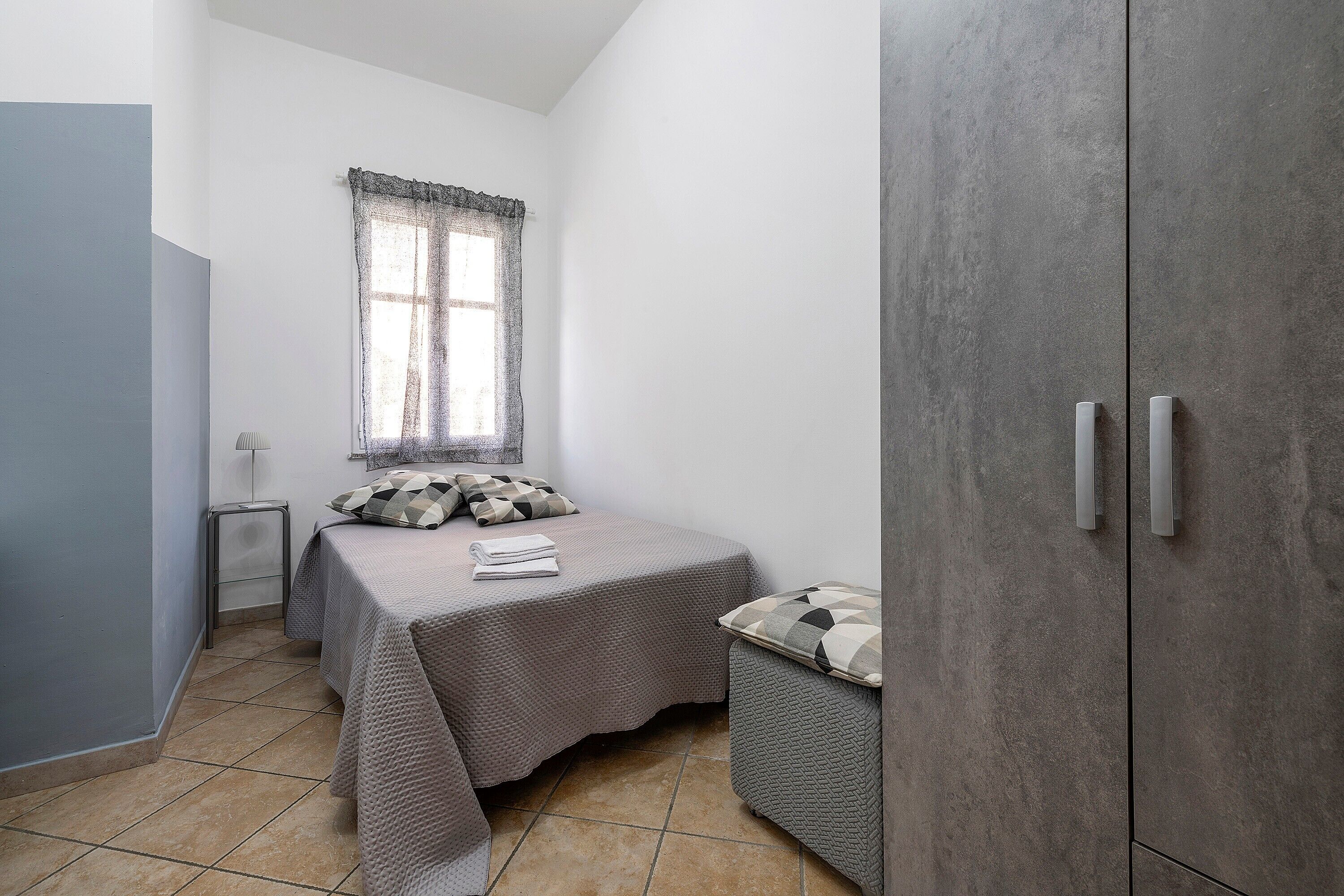 A bedroom with a bed, a window with lace curtains, a bedside table, and a wardrobe.