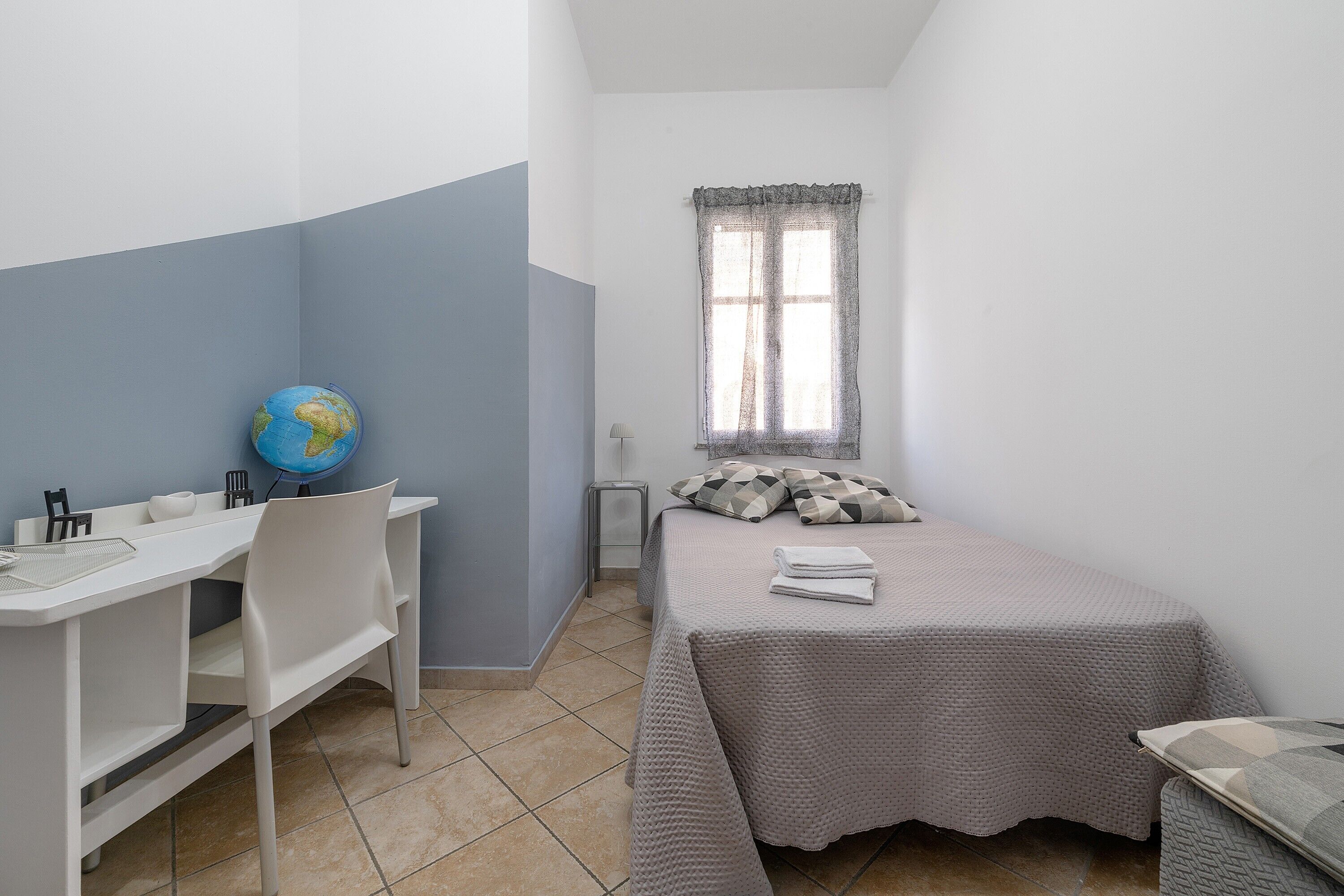 A small, neatly arranged bedroom with a desk, chair, bed, and a window with curtains.