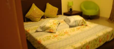 A bed with floral bedding, a nightstand with a lamp, and a green chair in a room with a brown headboard.