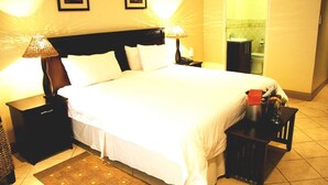 Una camera d'albergo con un letto grande, due lampade ai comodini, un tavolino notte e un piccolo tavolo con un oggetto rosso, oltre a un bagno visibile attraverso una porta aperta.