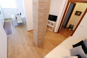 Ein Wohnzimmer mit einem weißen Sofa, einem Fernseher auf einem Lowboard, einem Essbereich mit Tisch und Stühlen sowie einer Wand mit Steinverkleidung.