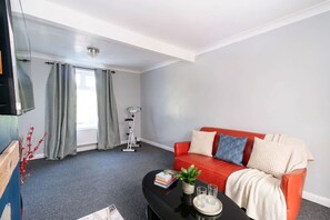 A living room with a red sofa, a coffee table with books and a plant, and a window with curtains. - Comfortable 3 Bed House nr Chesterfield w Garden (Doe Lea)
