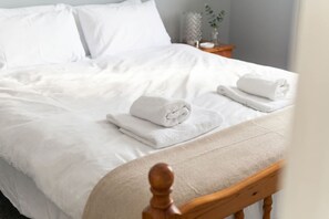 A neatly made bed with white linens and folded towels, a wooden headboard, and a small bedside table with a vase.