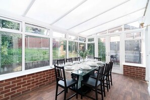 A conservatory with a glass table set for dining, surrounded by chairs, and large windows.