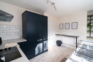 Une cuisine moderne avec des meubles de couleur sombre, un plan de travail blanc et une cuisinière noire. Une fenêtre avec des stores, un radiateur et une petite plante se trouvent sur le côté droit.
