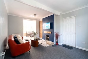 A living room with a red leather sofa, a flat-screen TV mounted on the wall, a fireplace, and a door.