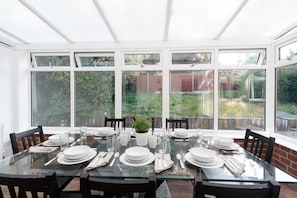 A conservatory dining area with a glass table set for six, surrounded by large windows offering a view of the garden. - Comfortable 3 Bed House nr Chesterfield w Garden (Doe Lea)